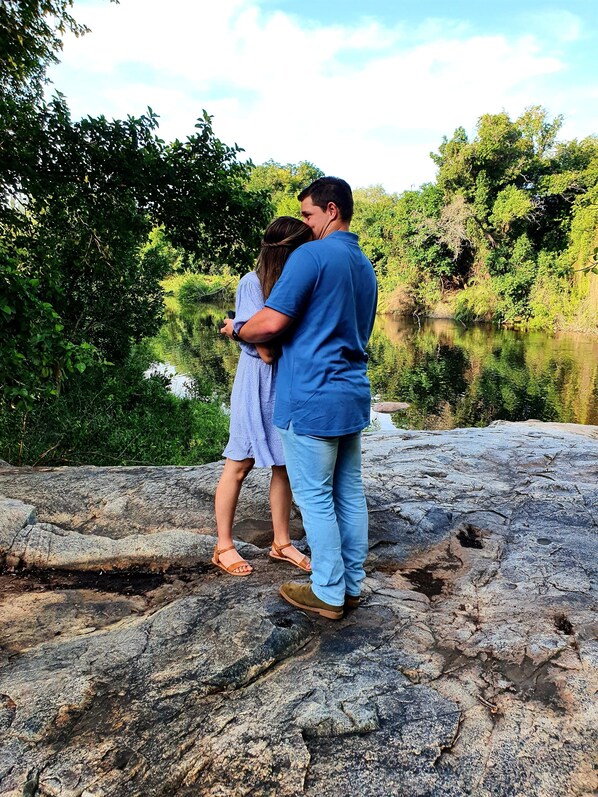 A couple standing on a rocky surface by a calm body of water, surrounded by lush greenery. - Blyde River Wilderness Lodge (Hoedspruit)