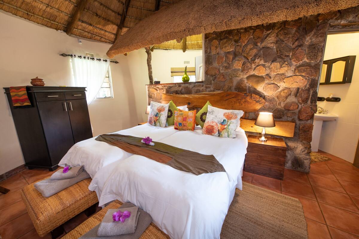 A well-appointed bedroom with a stone wall, a bed with floral pillows, a nightstand with a lamp, and a wooden bench with towels.