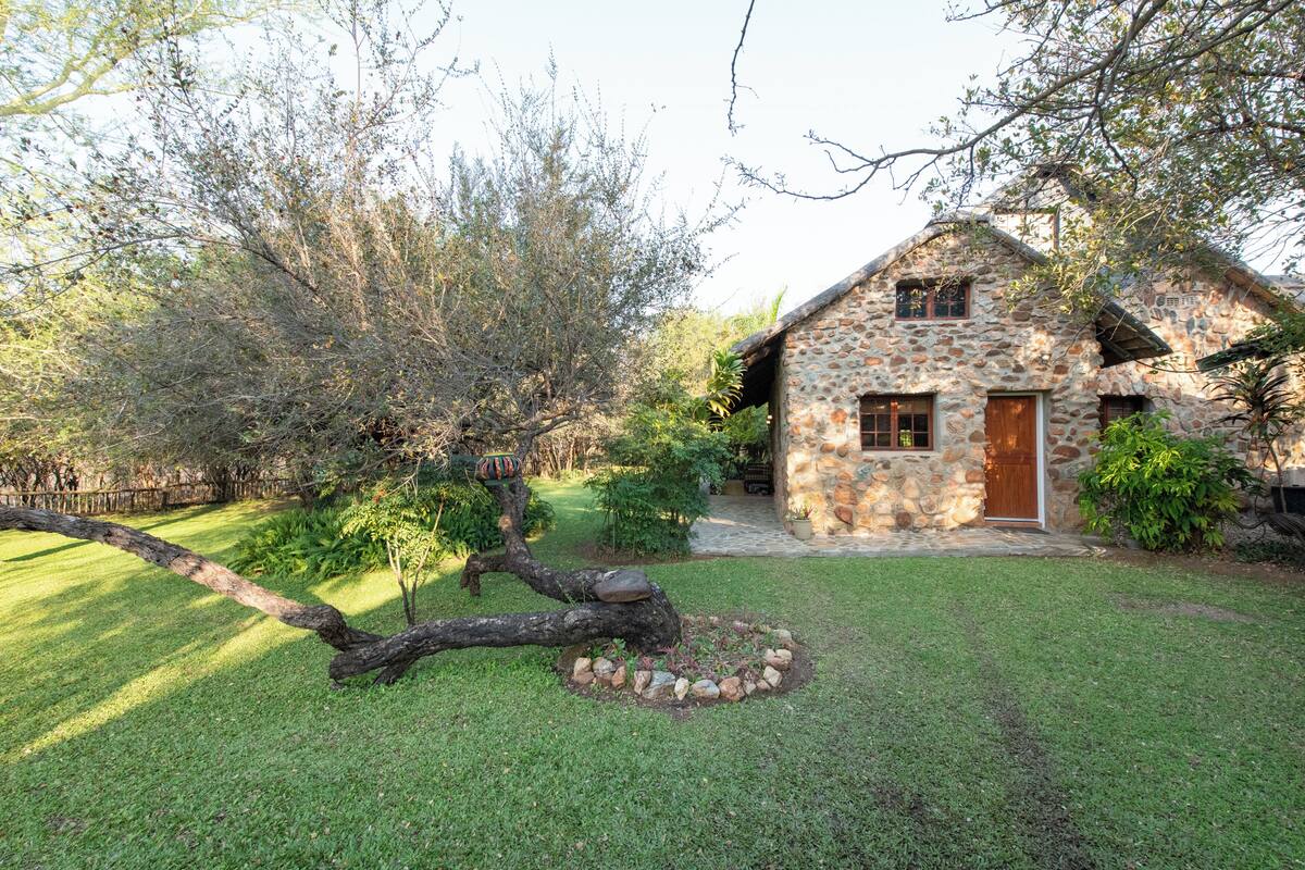 A stone house with a wooden door and windows, surrounded by a well-maintained lawn and trees.