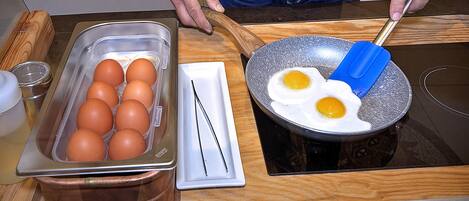 Una encimera de cocina con una bandeja de huevos, una sartén con dos huevos y una persona cocinando.