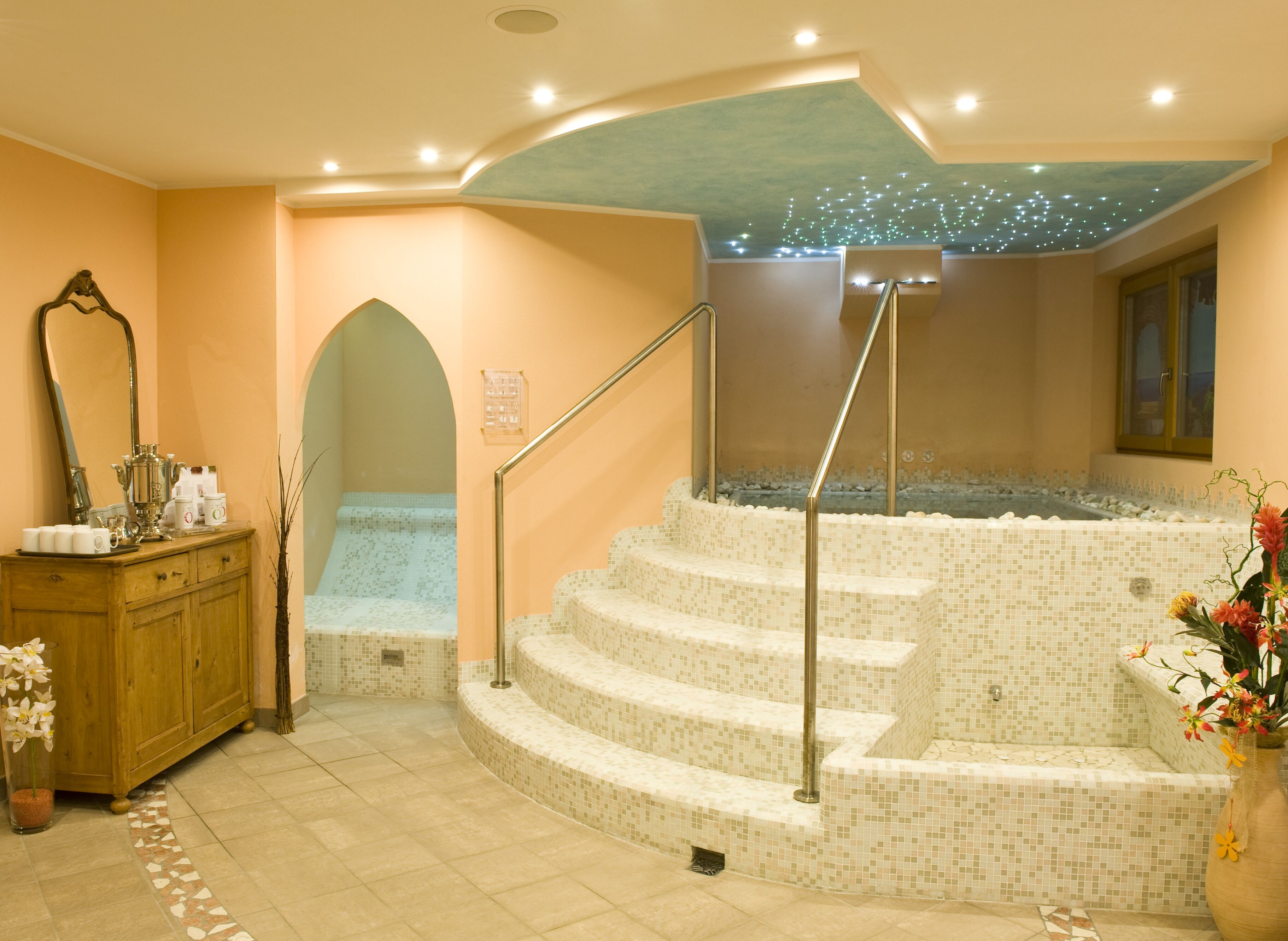Un espacio de spa con un jacuzzi revestido de mosaicos, un tocador de madera con artículos de aseo y un espejo.