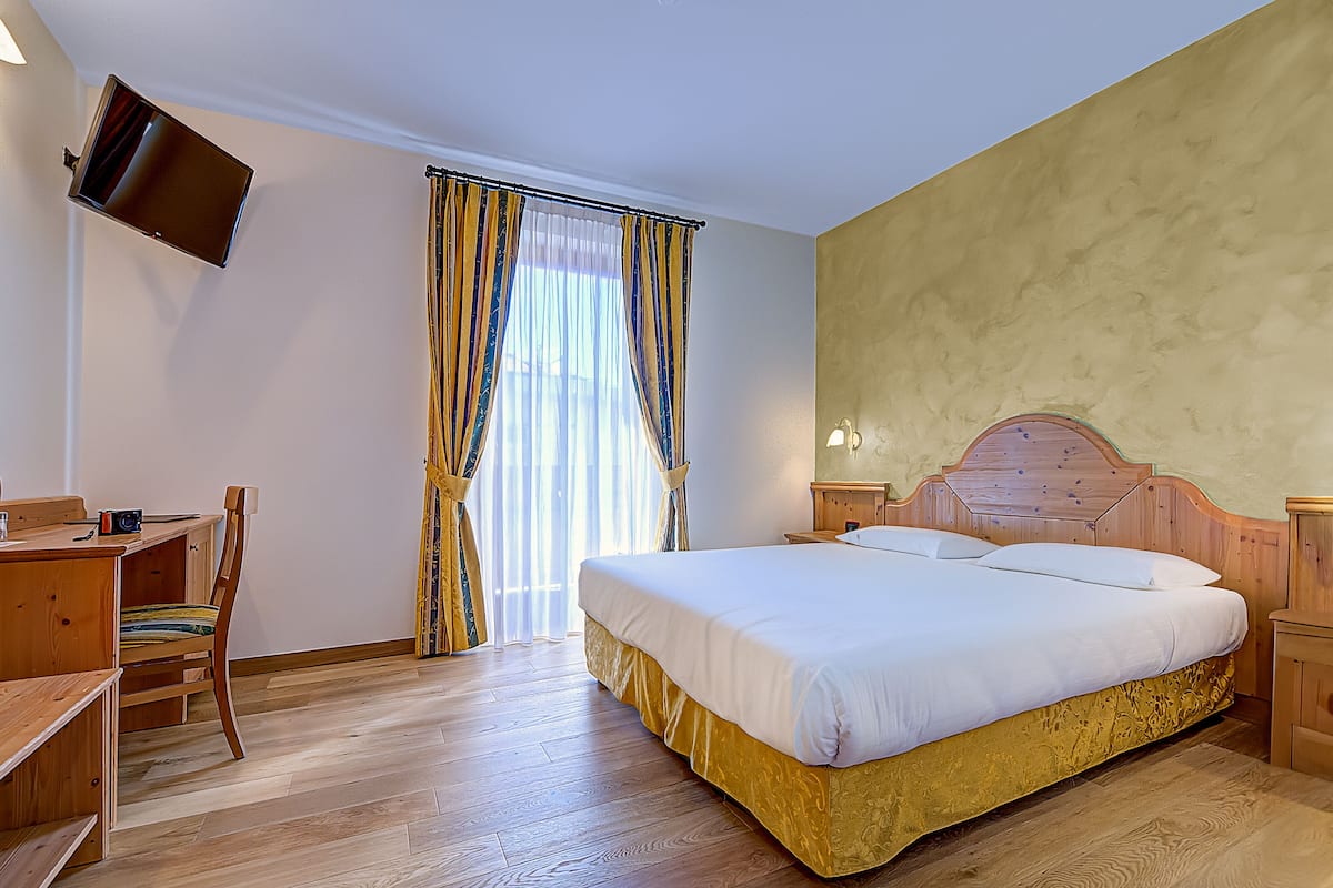 A hotel room with a wooden bed, a desk with a chair, a television mounted on the wall, and a window with curtains.