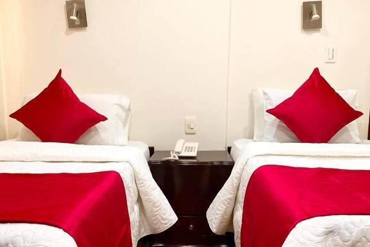 Een hotelkamer met twee bedden, elk met een rood kussen en witte beddengoed. Er staat een nachtkastje met een telefoon tussen de bedden.