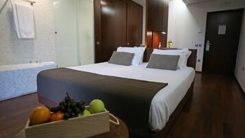 A hotel room with a large bed, a wooden headboard, a bedside table with a fruit basket, a bathroom with a towel hanging, and a door with a sign.