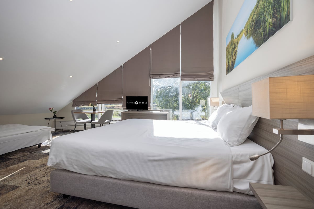 Quarto de hotel moderno com uma cama grande, uma escrivaninha e uma cadeira. Há um quadro na parede e uma janela com vista para o exterior.