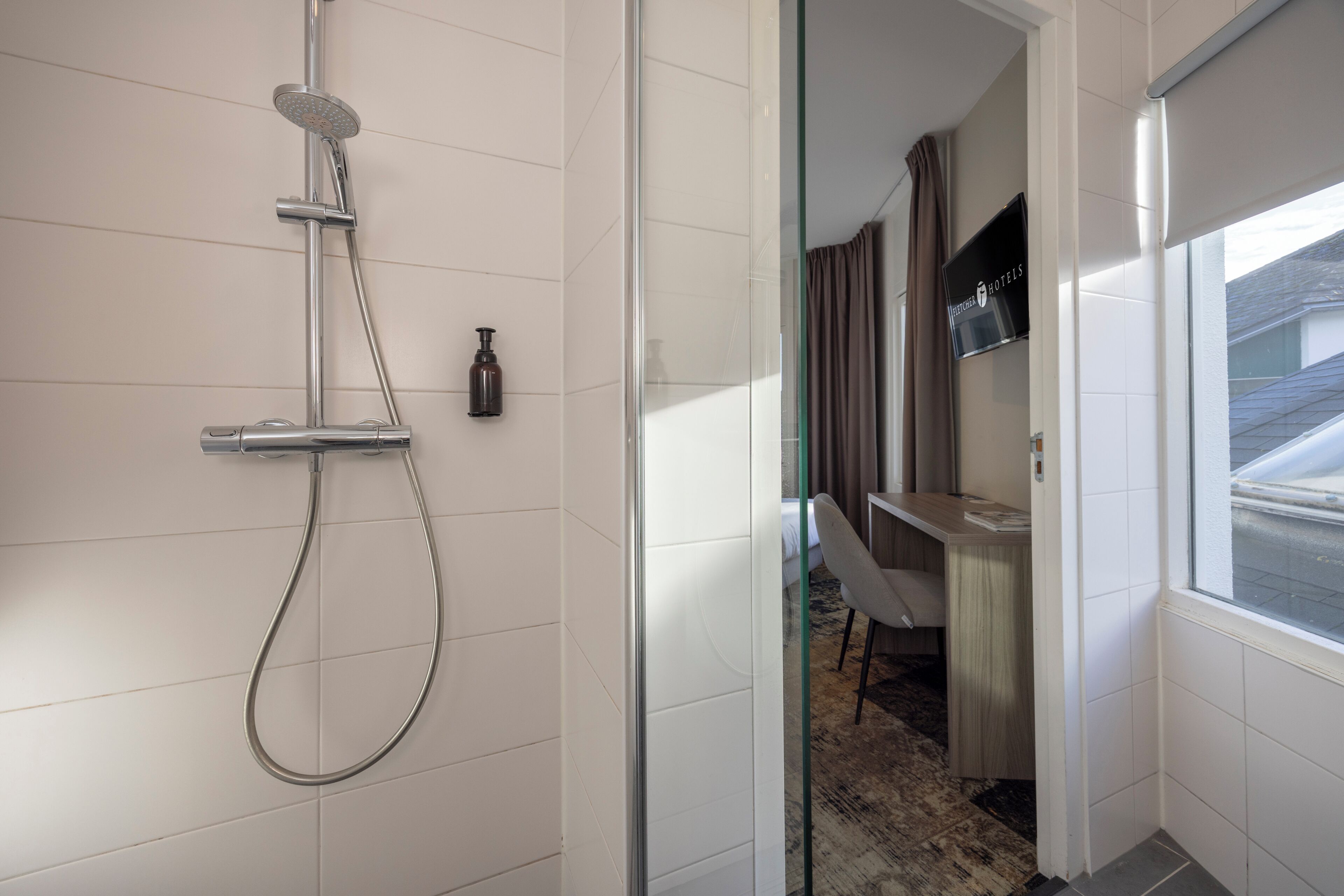Un baño moderno con ducha, inodoro y lavamanos. Una puerta de cristal da a una habitación con escritorio y silla.