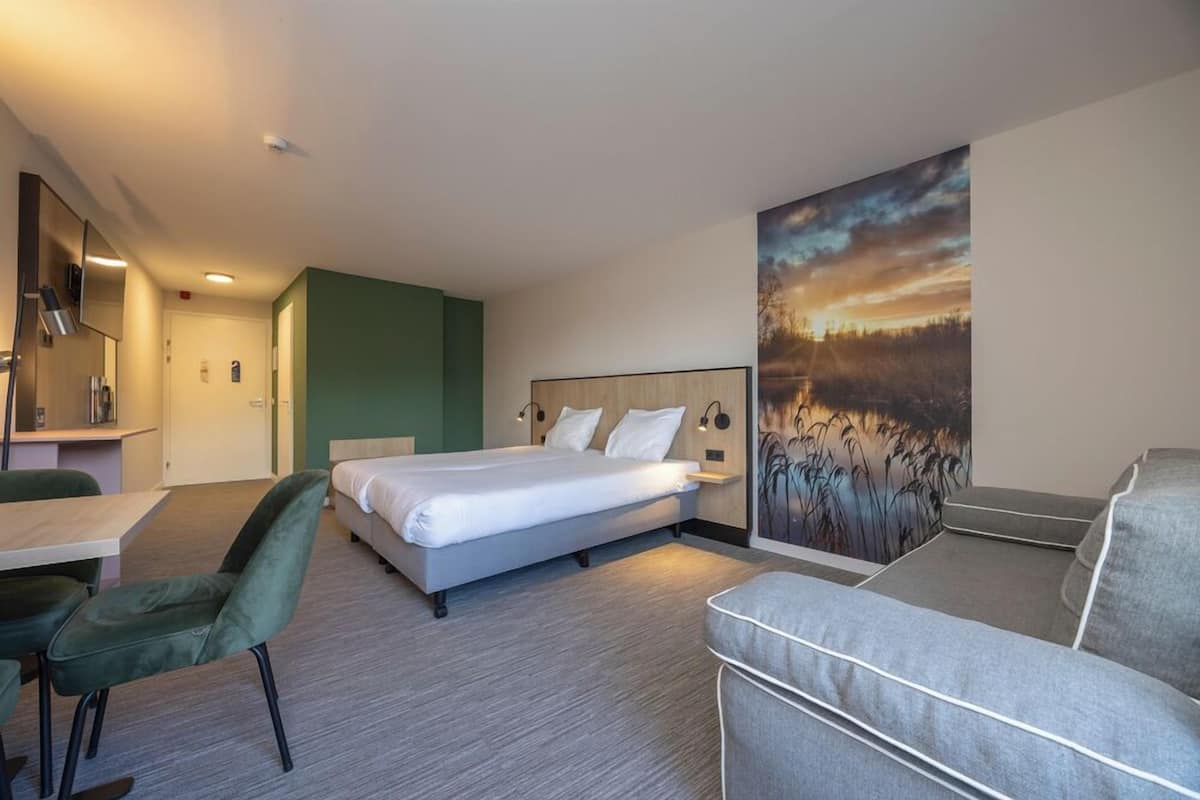 Quarto de hotel com cama, escrivaninha, poltrona, sofá e um quadro na parede.