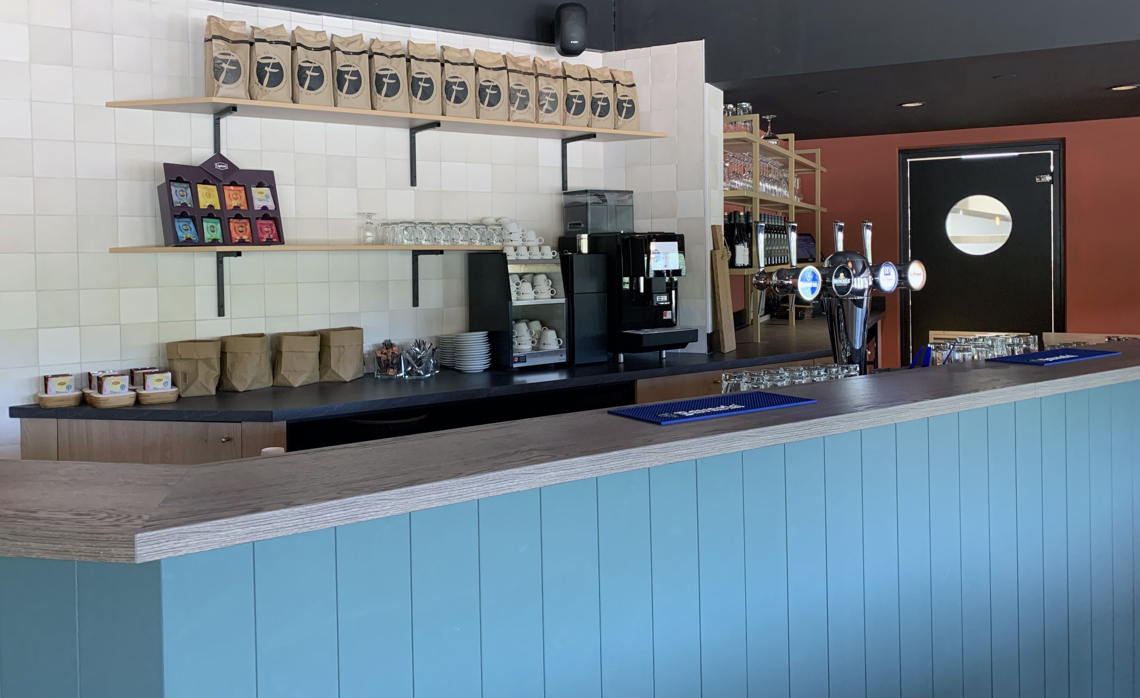 Mostrador de cafetería con una máquina de café, un grifo de cerveza y una estantería con sacos de café.