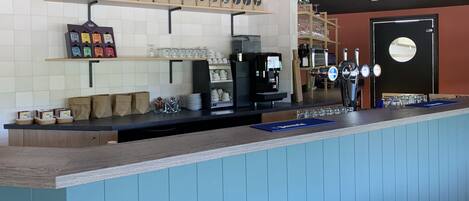 Mostrador de cafetería con una máquina de café, un grifo de cerveza y una estantería con sacos de café.