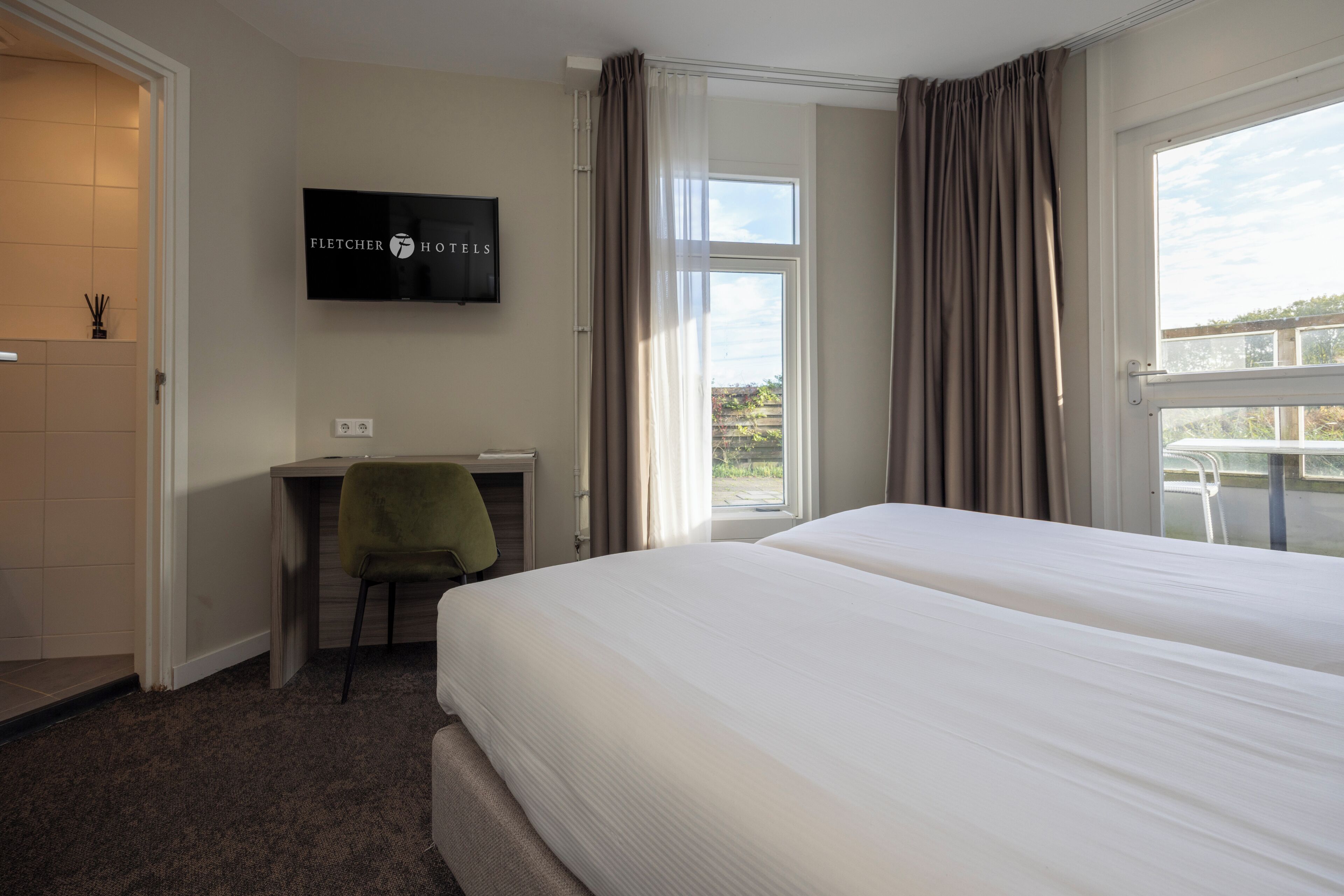 Habitación de hotel con cama, escritorio, silla, televisor y ventana con cortinas.