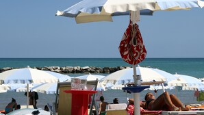 A beach with multiple umbrellas providing shade, a wooden table with a red top, and people relaxing by the water. - Residence I Delfini (Cupra Marittima)