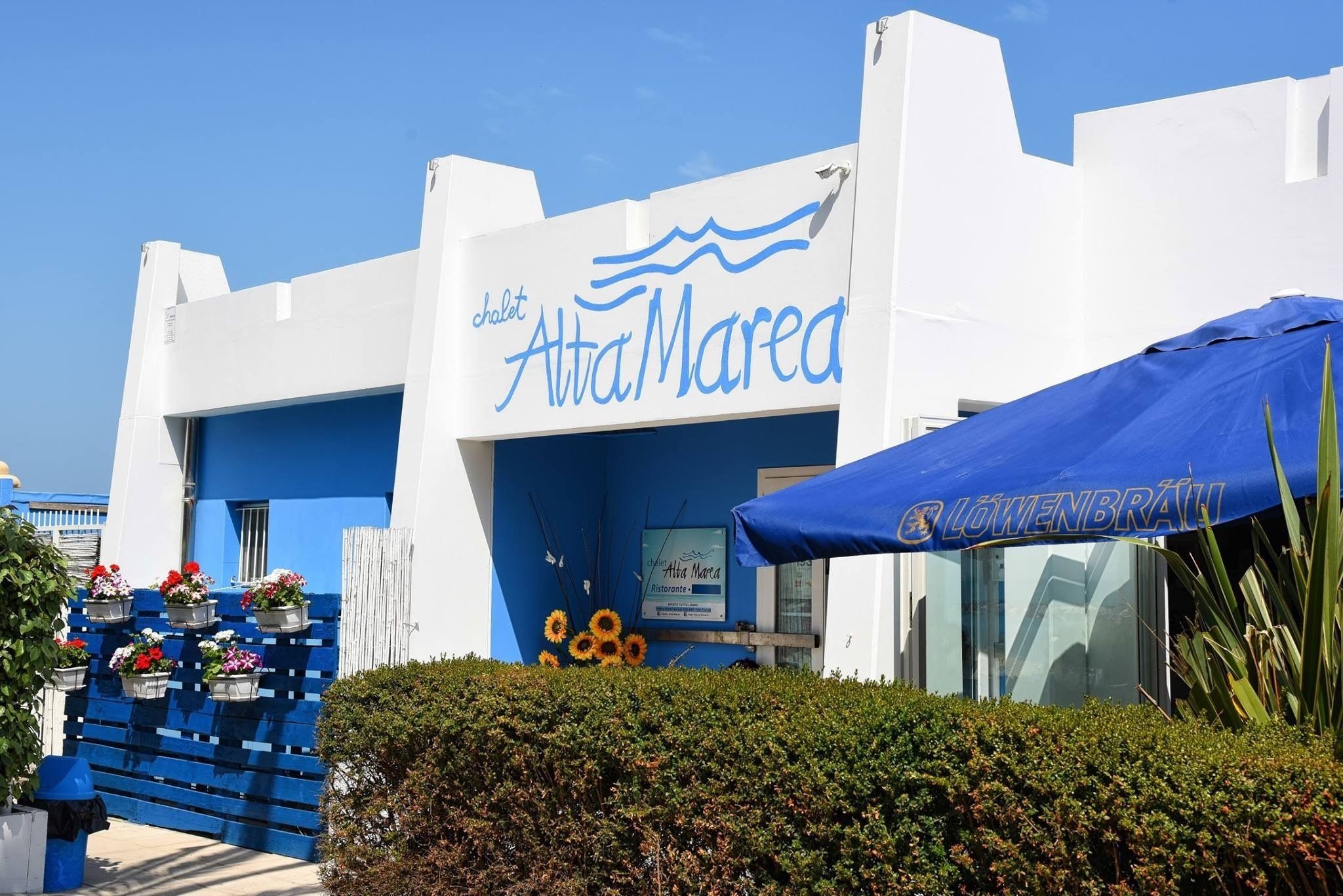 A building with a blue and white facade, a sign reading "chalet Alta Marea," and a blue awning with "Löwenbräu" written on it.