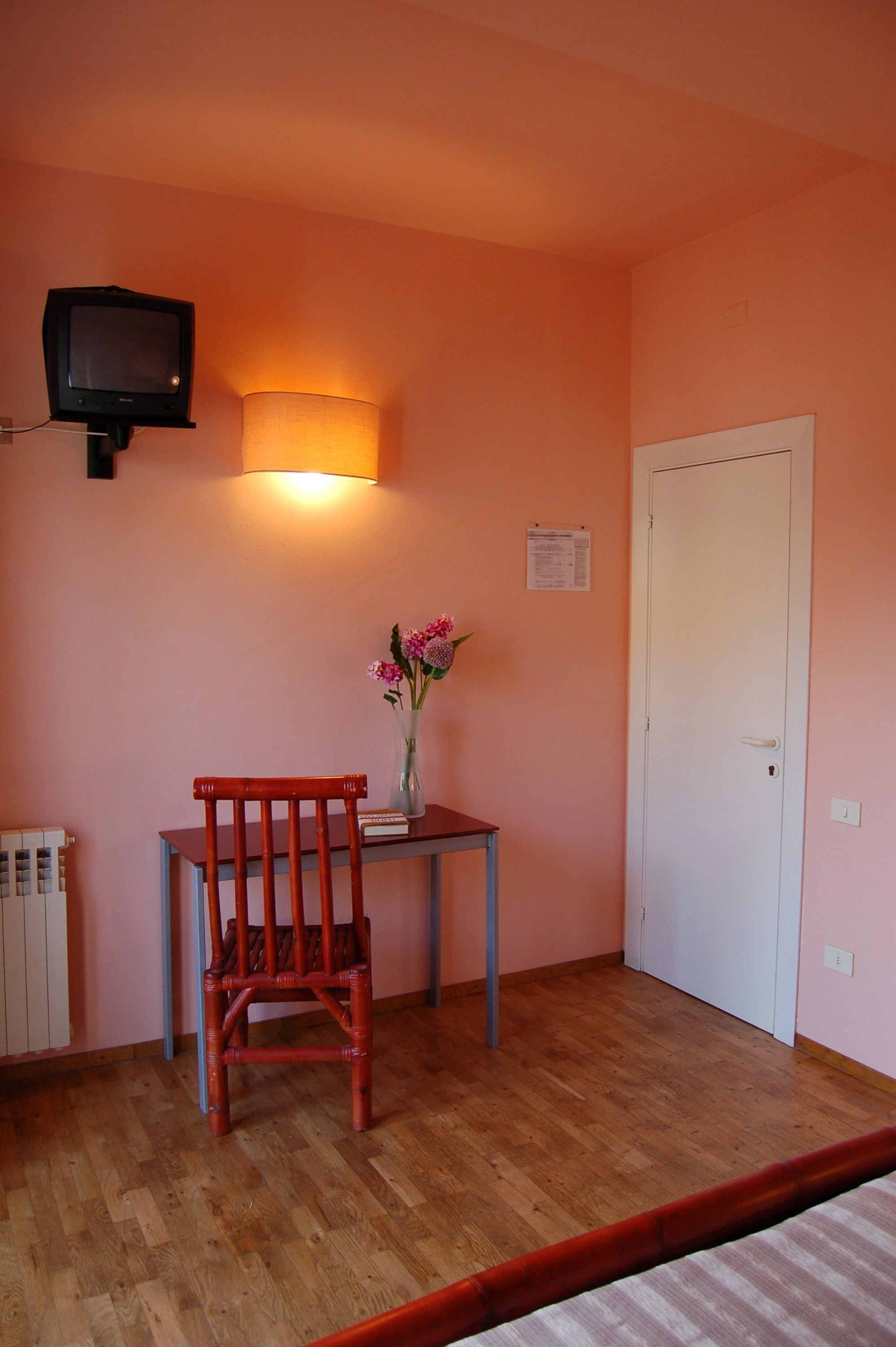 Una habitación con piso de madera, una silla roja, una pequeña mesa con un florero y una televisión montada en la pared, además de una puerta blanca.