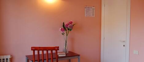 Una habitación con piso de madera, una silla roja, una pequeña mesa con un florero y una televisión montada en la pared, además de una puerta blanca.