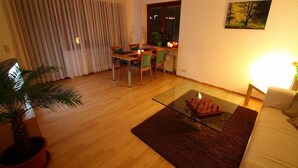 Un salon avec une table basse en verre, un canapé, une table à manger avec des chaises, une plante en pot et un tableau accroché au mur.