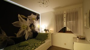 Une chambre à coucher avec un lit, une lampe fixée au mur, une fenêtre avec des rideaux et une petite table sur laquelle est posée une plante.