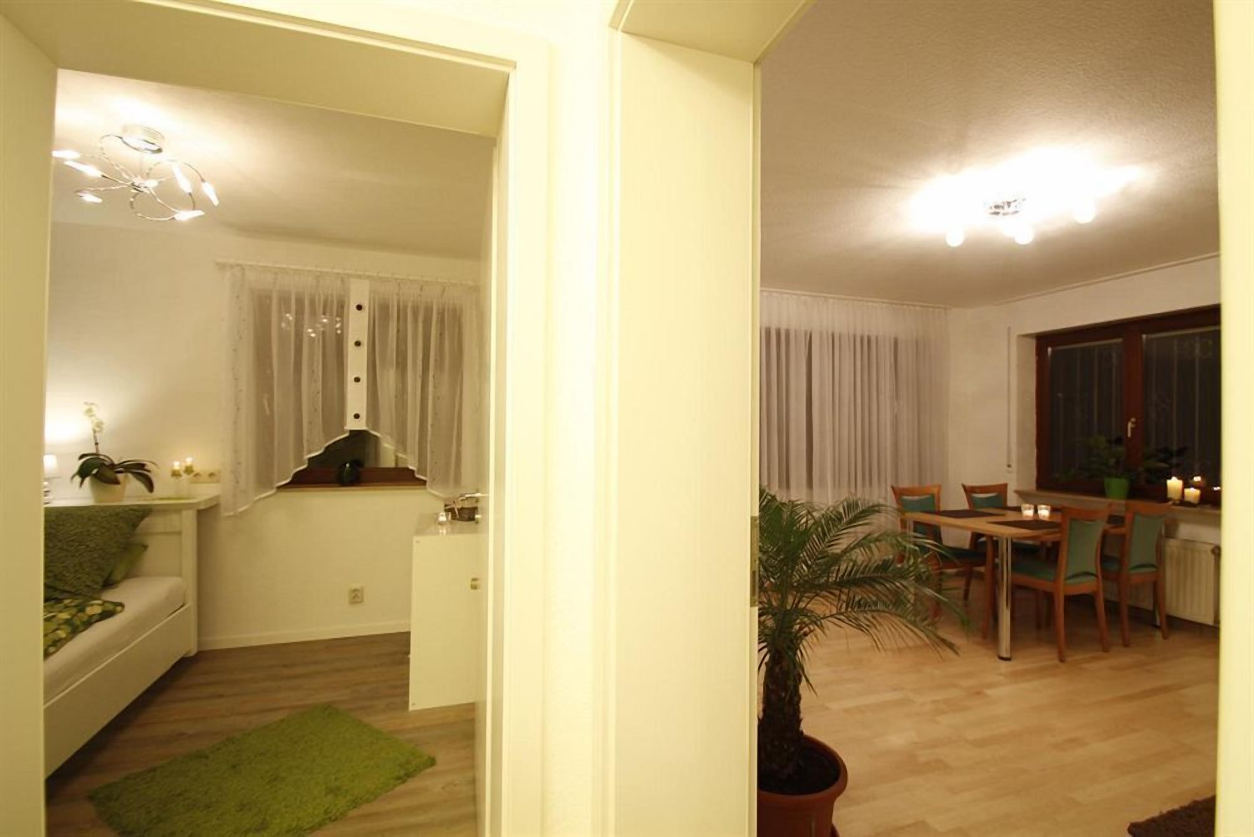 Un salón luminoso con sofá, una mesa de comedor con sillas, una planta en maceta y una pequeña cocina con ventana.