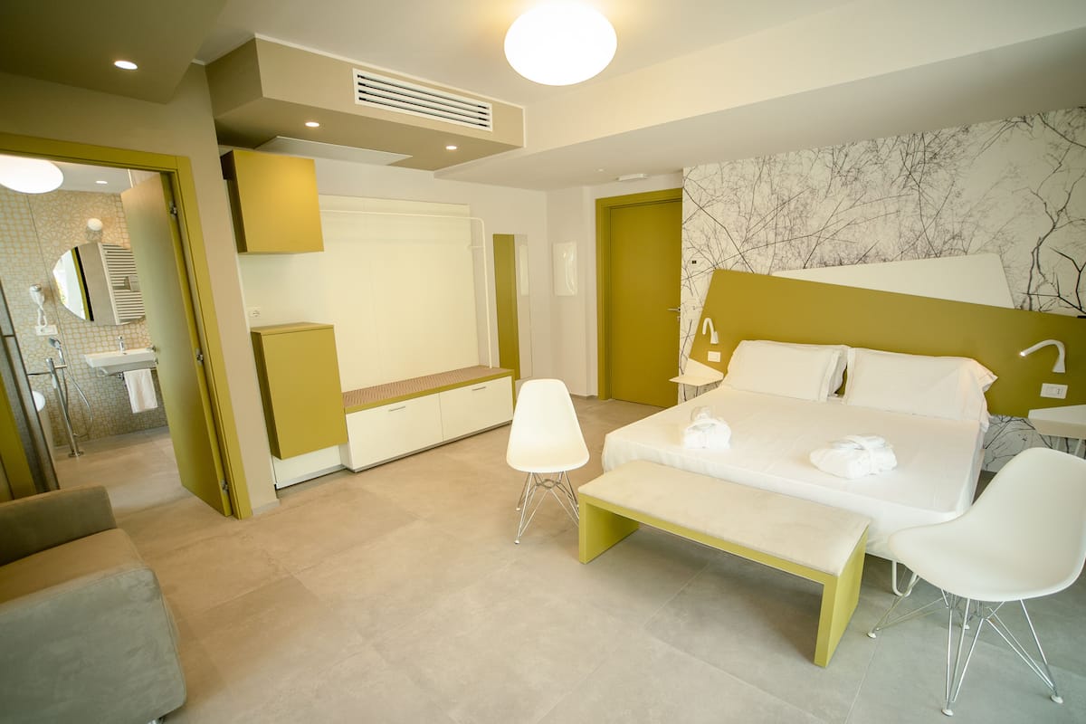 A modern hotel room with a bed, bedside table, two chairs, a bench, a bathroom with a mirror, sink, and toilet.