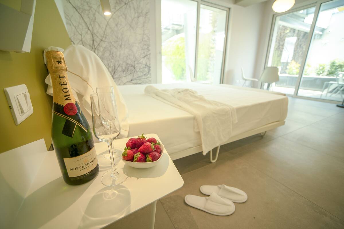 A hotel room with a bed, a bottle of champagne, a bowl of strawberries, and a pair of slippers on the floor.