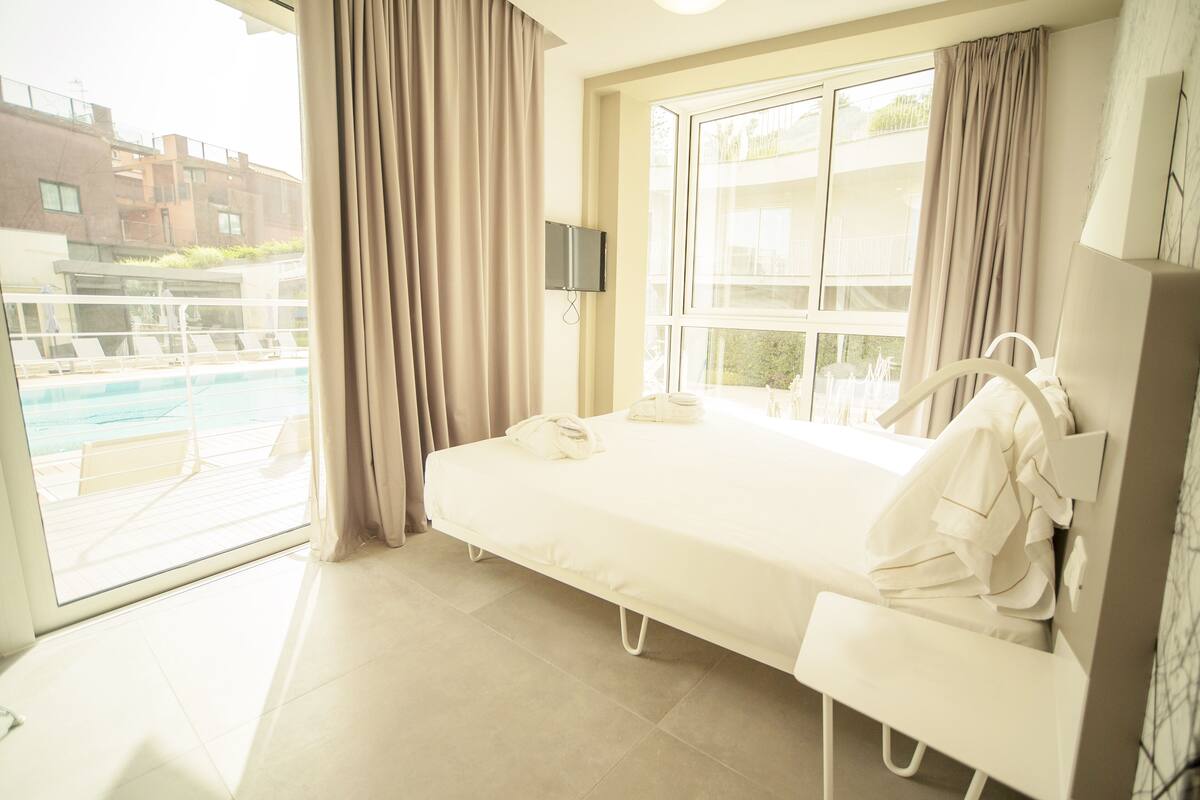 A hotel room with a bed, a TV, a window with curtains, and a view of a pool and buildings outside.