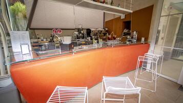 Zona de bar moderna con una barra curva color naranja, sillas de metal blanco y diversos equipos de bar.