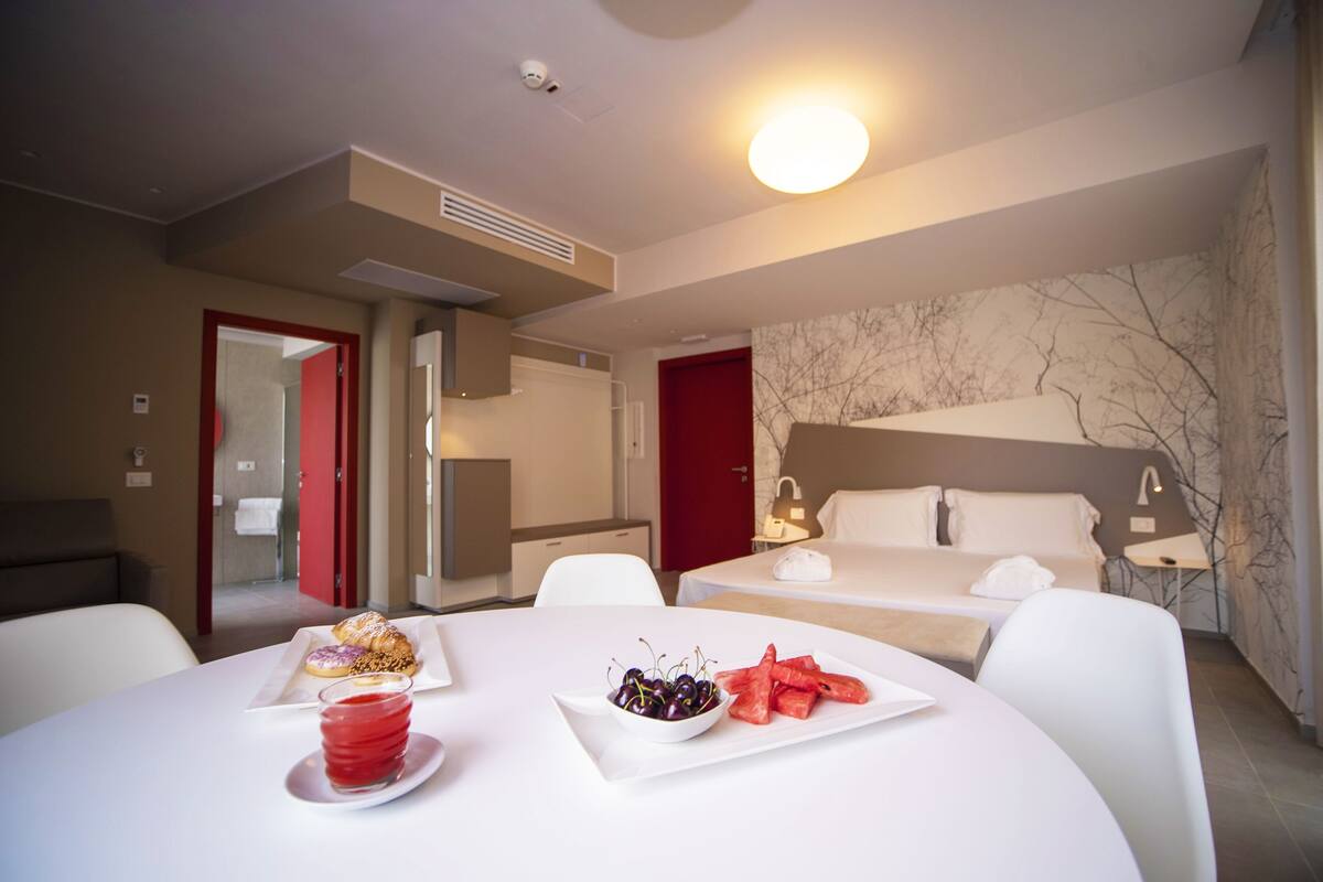 A hotel room with a bed, a bedside table, a chair, a dining table with food, and a bathroom visible through an open door.