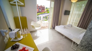 Habitación de hotel con cama, una mesita con un plato de fruta, una silla y un balcón con vistas a edificios y zonas verdes.