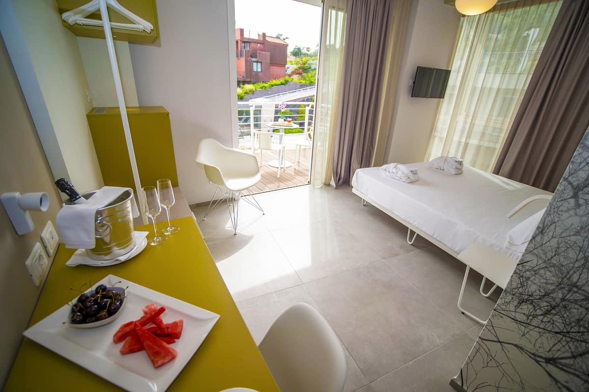 A hotel room with a bed, a small table with a plate of fruit, a chair, and a balcony with a view of buildings and greenery.