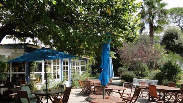 Eine Terrasse im Freien mit Holztischen und -stühlen, einem blauen Sonnenschirm und einer weißen Laube.