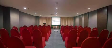 Un salón de conferencias con filas de sillas rojas, un atril y una pantalla de proyección que muestra