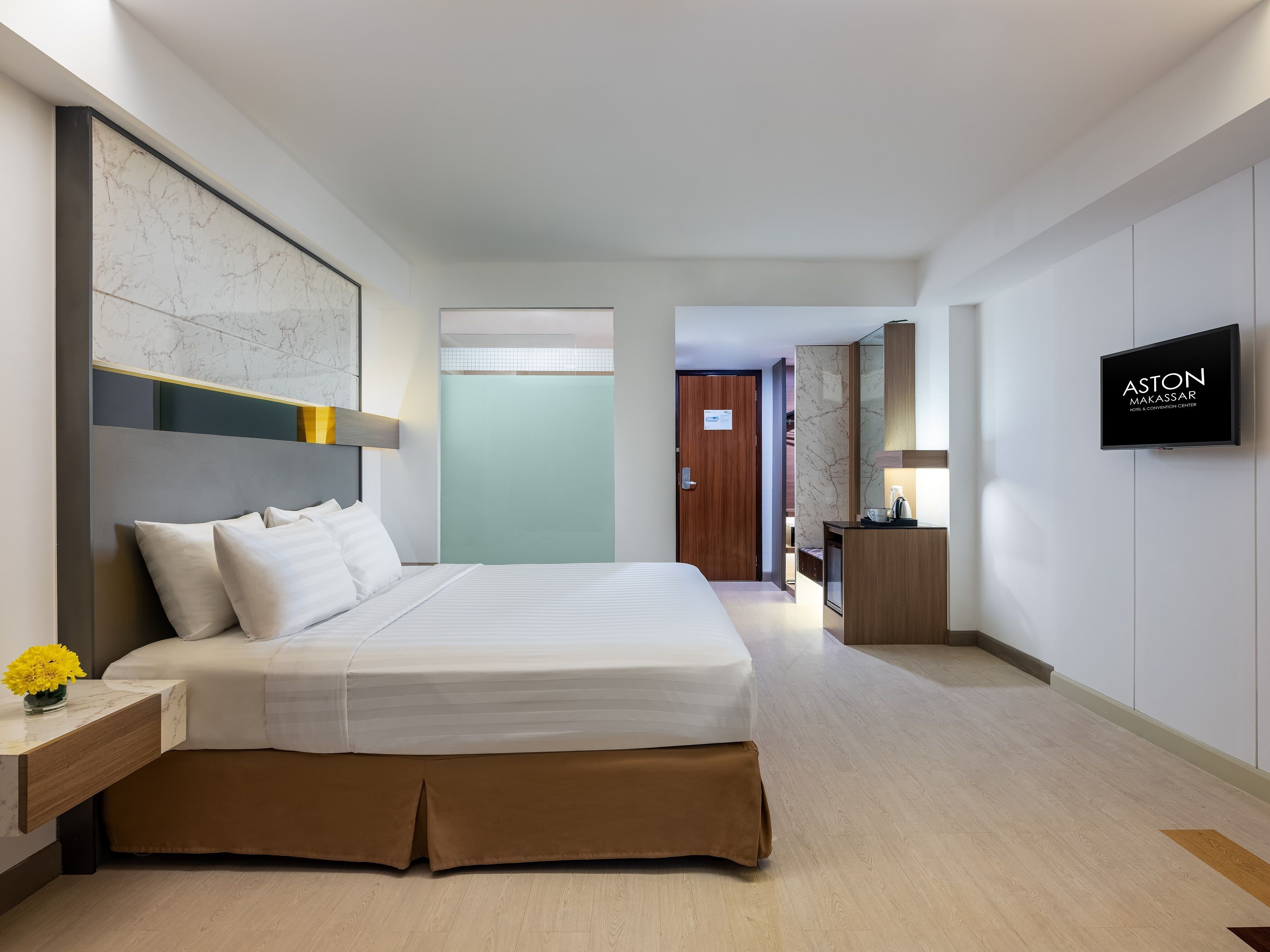 A modern hotel room with a large bed, a bedside table with a vase of flowers, a television, and a bathroom visible through an open door.