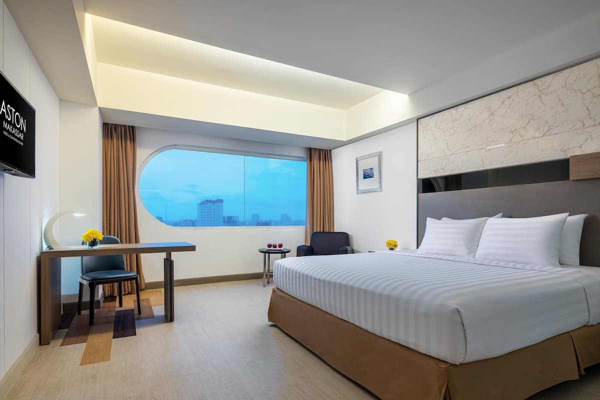 Una habitación de hotel moderna con una cama grande, un escritorio con una silla, una mesita con un florero y un ventanal con vistas a la ciudad.