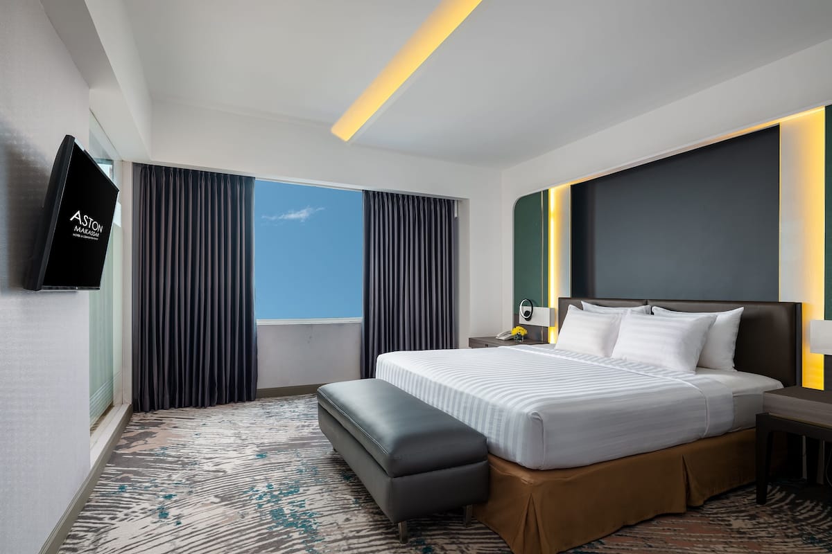Una habitación de hotel moderna con una cama grande, un televisor de pantalla plana, una mesita de noche con lámpara y vistas al cielo a través de la ventana.