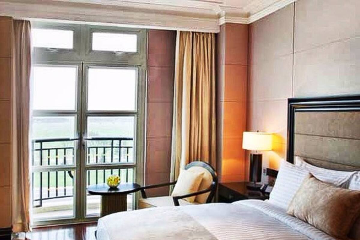 Habitación de hotel con cama, mesita de noche con lámpara, una silla, una pequeña mesa con un florero y un balcón con barandilla.