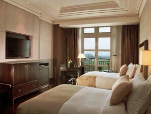 A hotel room with a large bed, a TV mounted on the wall, a desk with a lamp, and a view of a green landscape through the window.