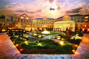 Un edificio grande e histórico con un patio central que tiene una fuente, rodeado de jardines e iluminados senderos.