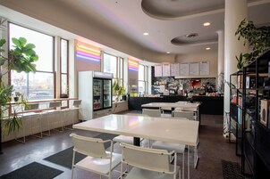 Un café moderno con mesas y sillas blancas, una zona de cocina y ventanas amplias con vistas a la ciudad.