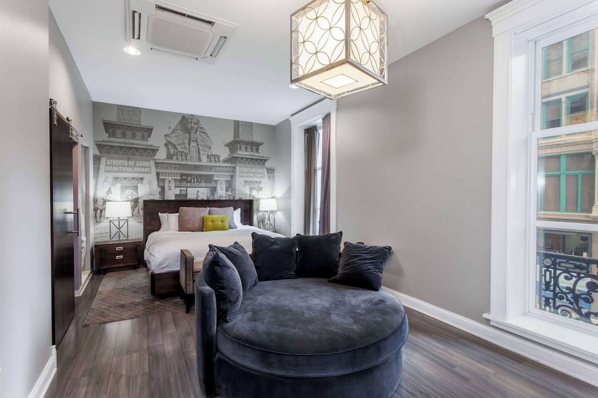 Une chambre d’hôtel moderne dotée d’un grand lit, d’un coin salon circulaire, d’une fresque représentant les rues du Caire et d’une grande fenêtre offrant une vue panoramique.