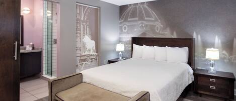 A bedroom with a large bed, a bedside table with a lamp, a wooden bench, and a glass shower enclosure.