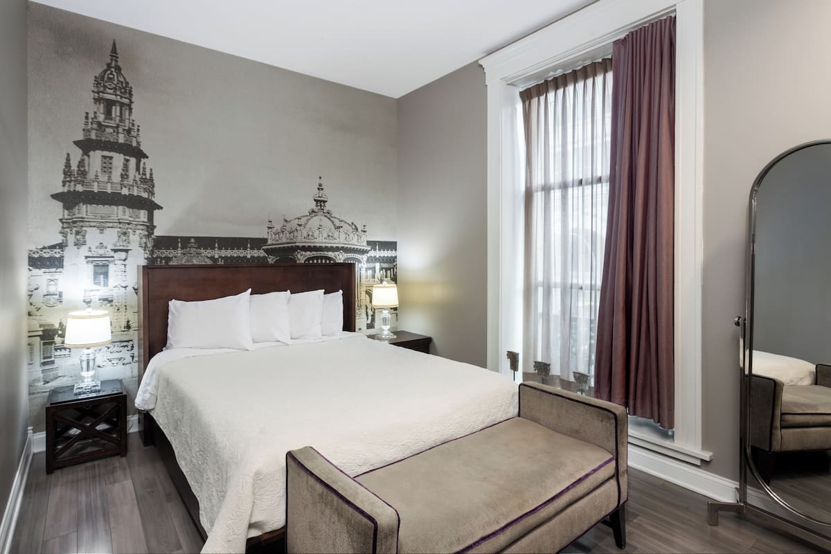 Une chambre d’hôtel avec un grand lit, une table de chevet avec une lampe, une chaise, un grand miroir et une fresque murale représentant un bâtiment.