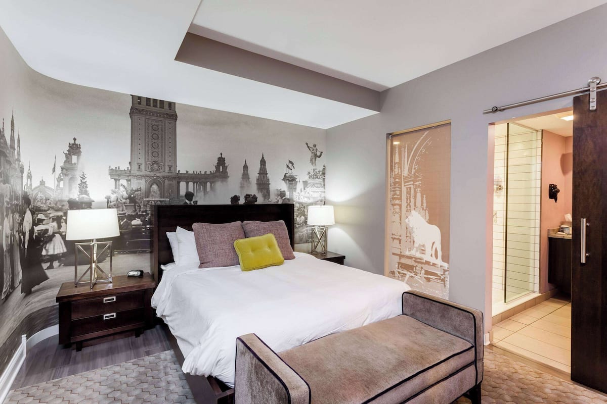 Une chambre à coucher avec un grand lit, des lampes de chevet, une table de chevet, un banc et une fresque représentant un paysage urbain.
