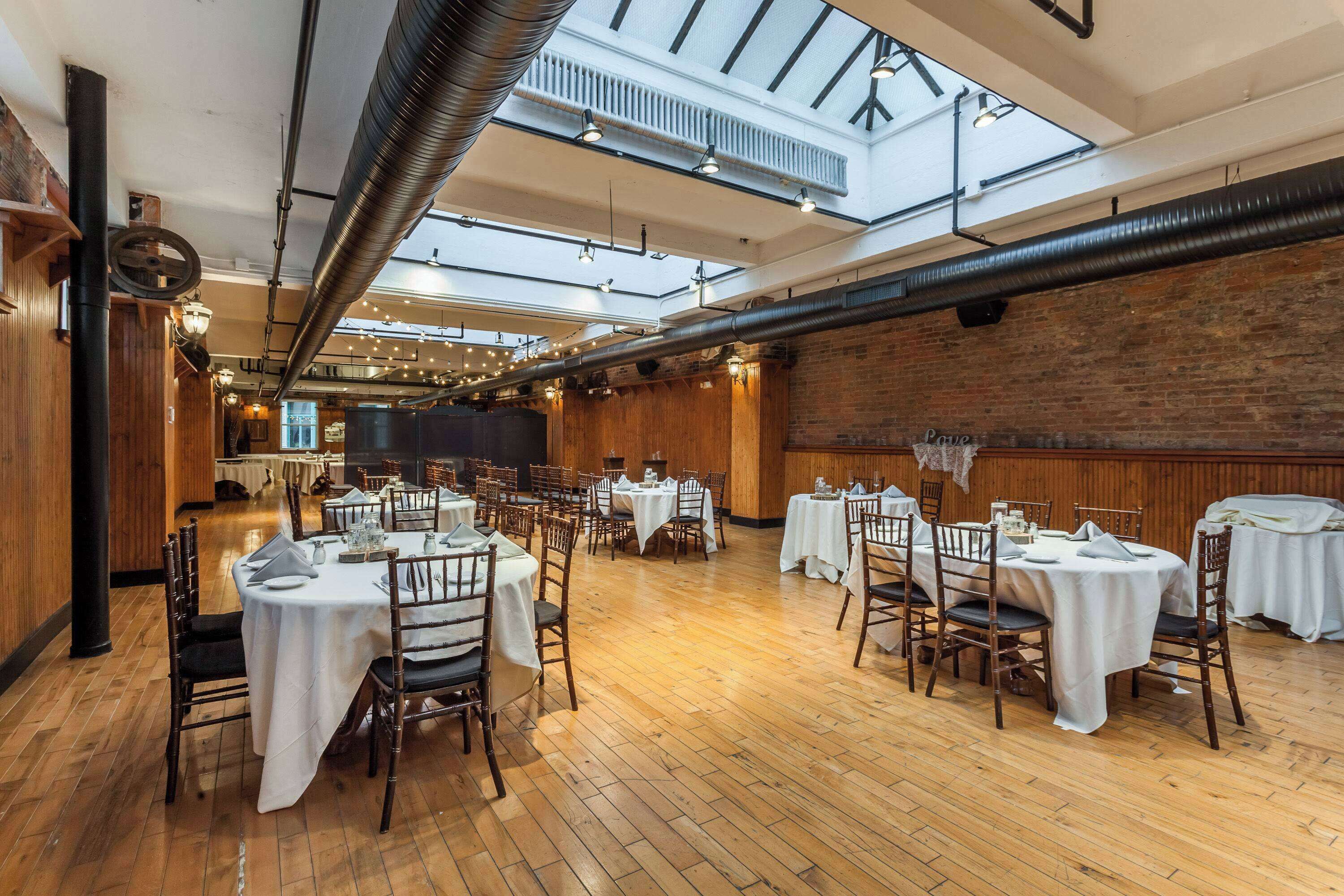 A spacious dining area with multiple tables and chairs, high ceilings with exposed ductwork, and a brick wall.