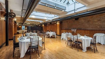 A spacious dining area with multiple tables and chairs, high ceilings with exposed ductwork, and a brick wall.