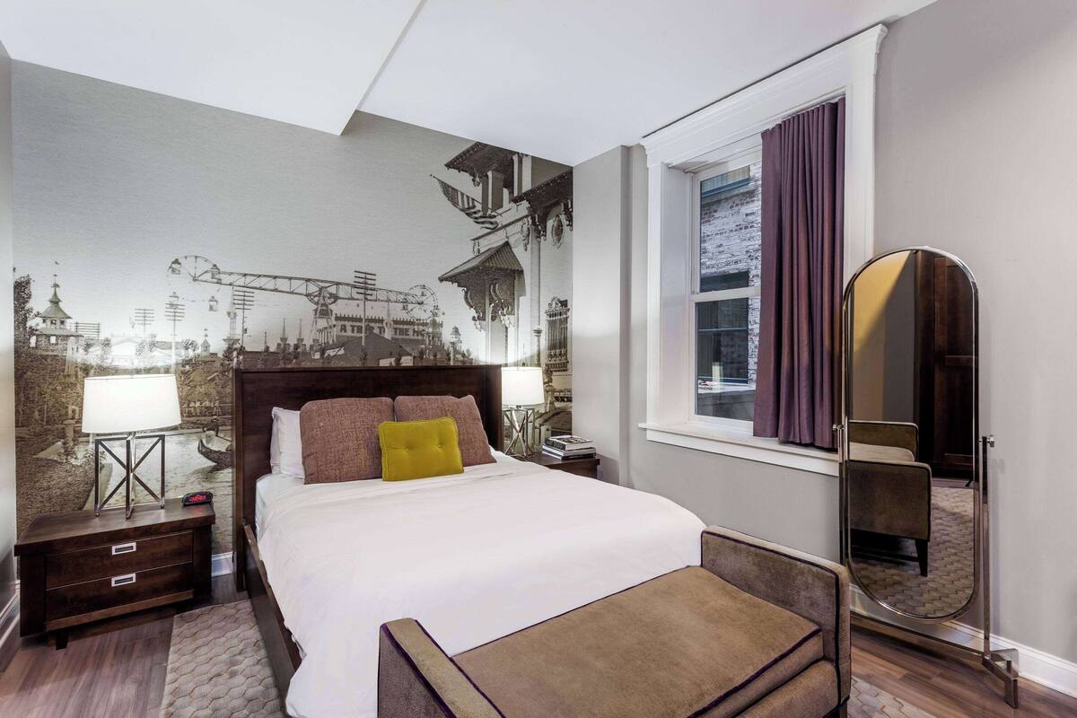 Une chambre d’hôtel avec un grand lit, une table de chevet, une commode, un miroir et une fenêtre avec des rideaux.