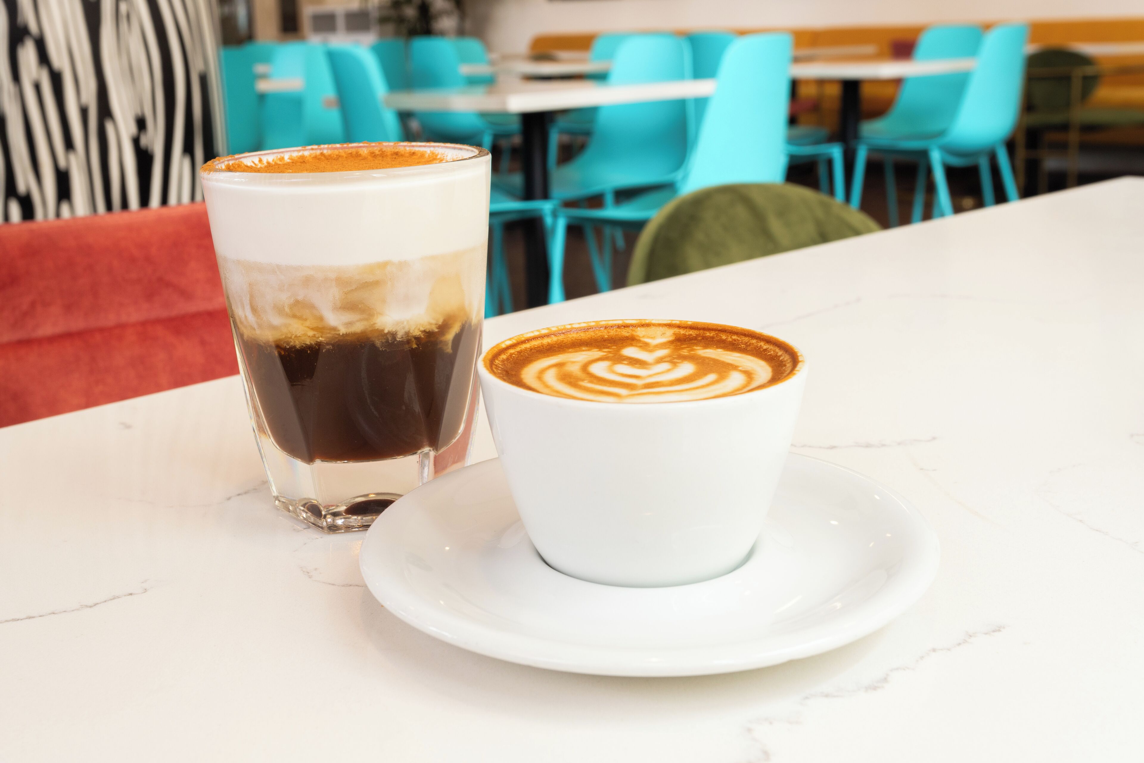 Un vaso y una taza de café sobre una mesa blanca en una cafetería, con sillas azules y un sofá rojo al fondo.