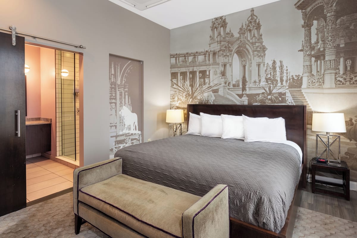 Une chambre à coucher avec un grand lit, deux tables de chevet avec des lampes, un banc, une porte coulissante et une fresque représentant un bâtiment.