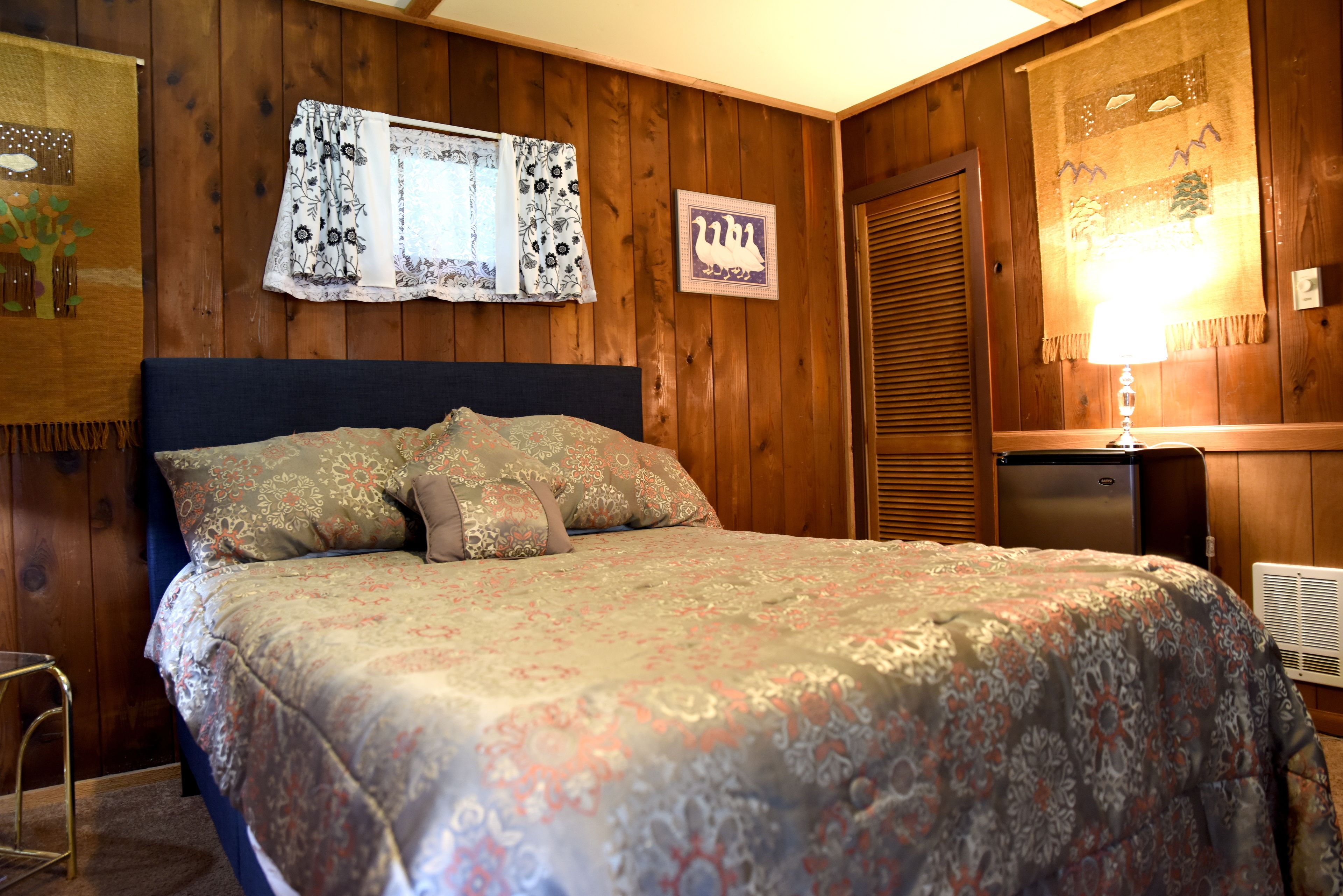 A bedroom with a large bed, wooden walls, a window with floral curtains, and a wall-mounted lamp.