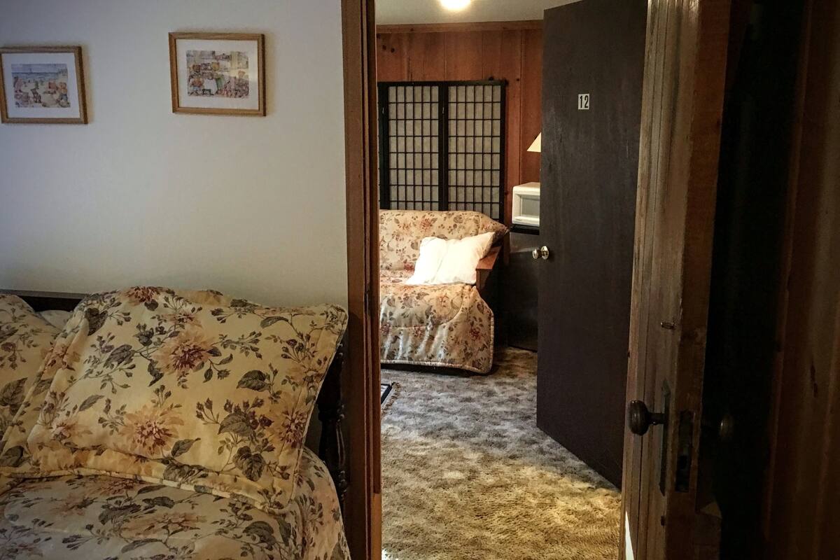 Quarto com cama de padrão floral, porta de madeira e um pequeno armário com micro-ondas.
