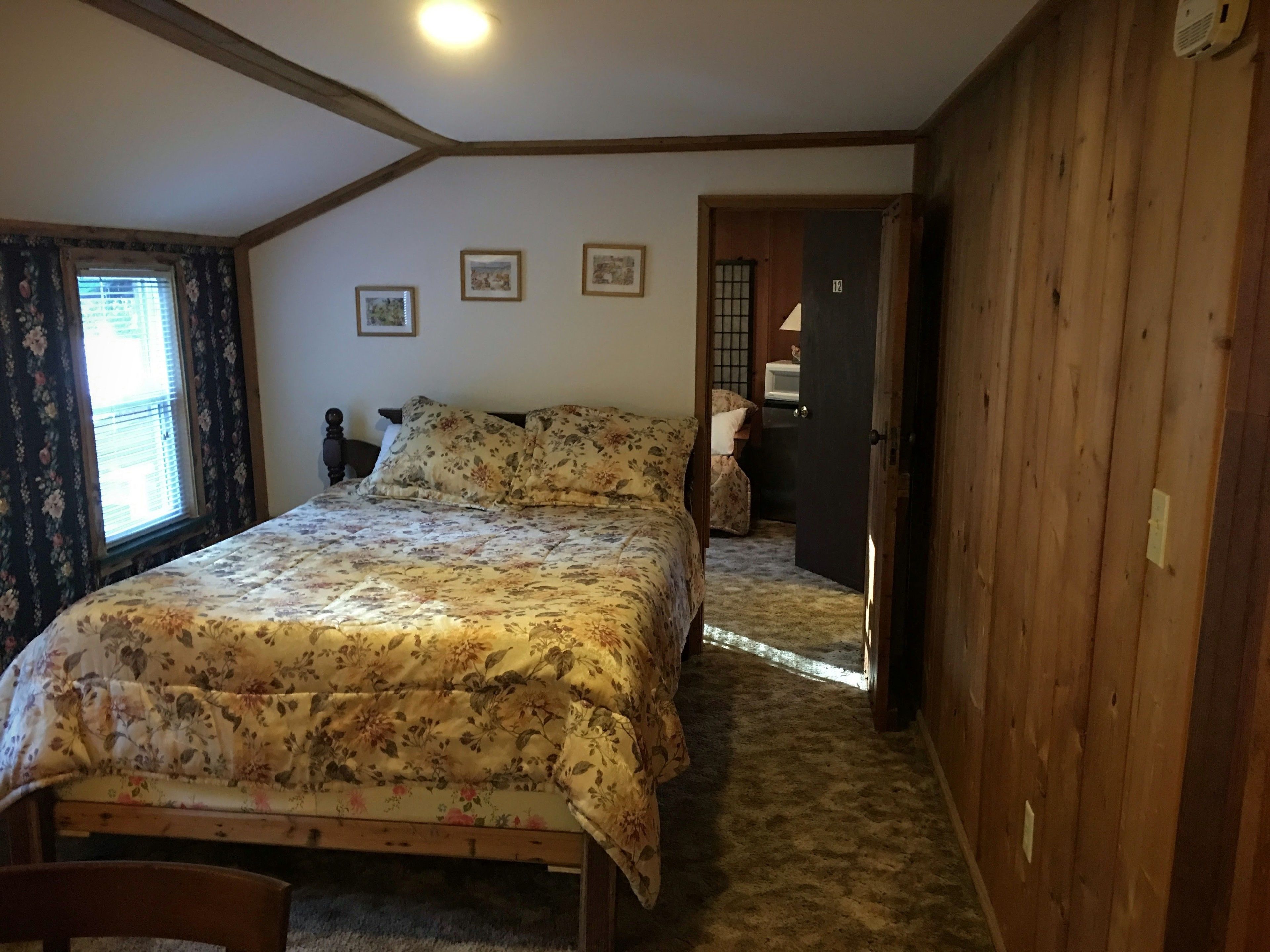 A bedroom with a wooden bed, a window with floral curtains, and a door leading to another room.