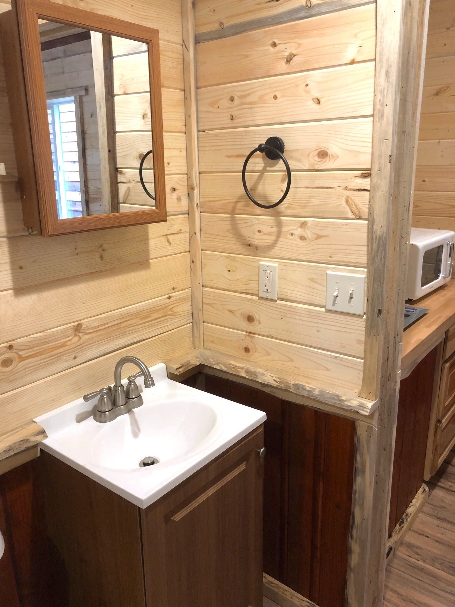 Baño de cabaña de madera con lavamanos blanco, espejo, aro para toallas y microondas.